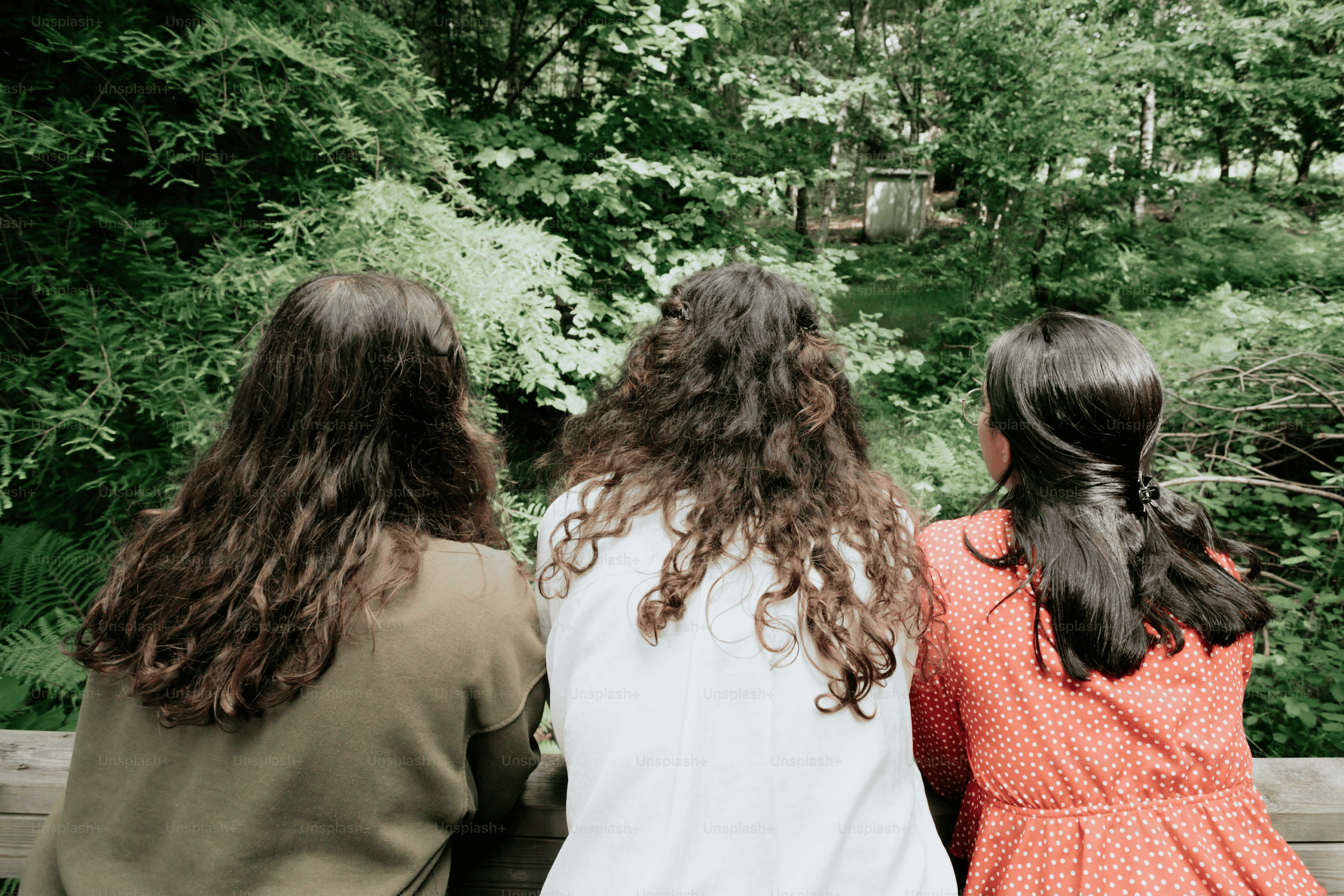 Three women sitting quietly together on a bench along a wooded path, reflecting shared presence and support during grief.